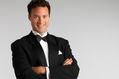 A smiling man in a tuxedo standing with his arms folded while looking into the camera.