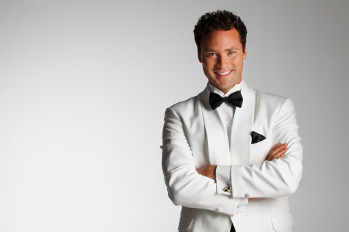 A smiling man in a white tuxedo standing with his arms folded while looking into the camera.