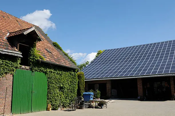 "Old farm building, partial covered with ivy, another farm building with a lot of solar panels in the background/ Germany."