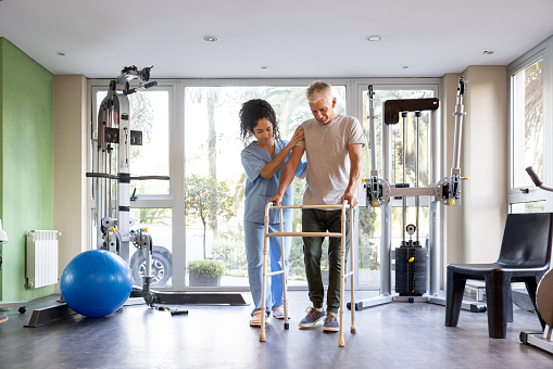 Mature man in physical therapy trying to walk using a walker with the help of his therapist - orthopedic medicine concepts