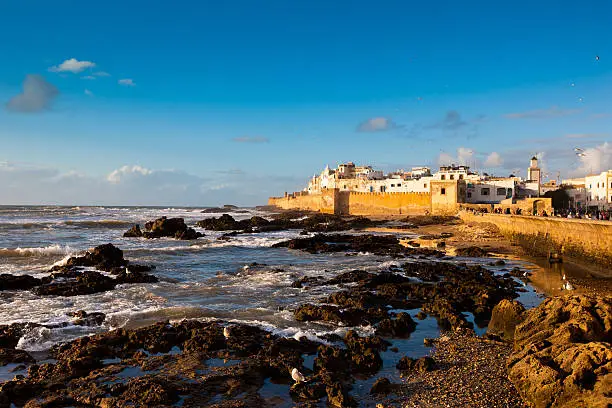 "Famous Port of Essaouira at the atlantic coast in morocco during sunset. Sun begins to illuminate the remparts of the old medina. Tourists and locals sitting together on the harbor walls enjoying the sunset. Essaouira, Morocco, North Africa."