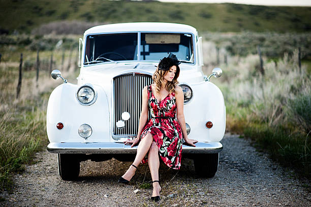 Beautiful young woman with vintage car Portrait of a beautiful woman sitting on the front bumper of a vintage car on a country dirt road. pin up girls and cars stock pictures, royalty-free photos & images