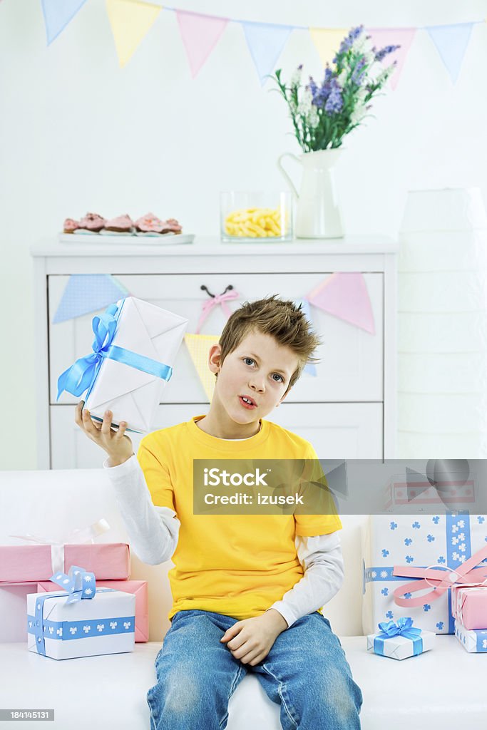 Cute boy with birthday present "Kids Birthday Party. Cute little boy sitting on sofa among many birthday presents and holding one of them in his hand, looking at camera and smiling." 8-9 Years Stock Photo