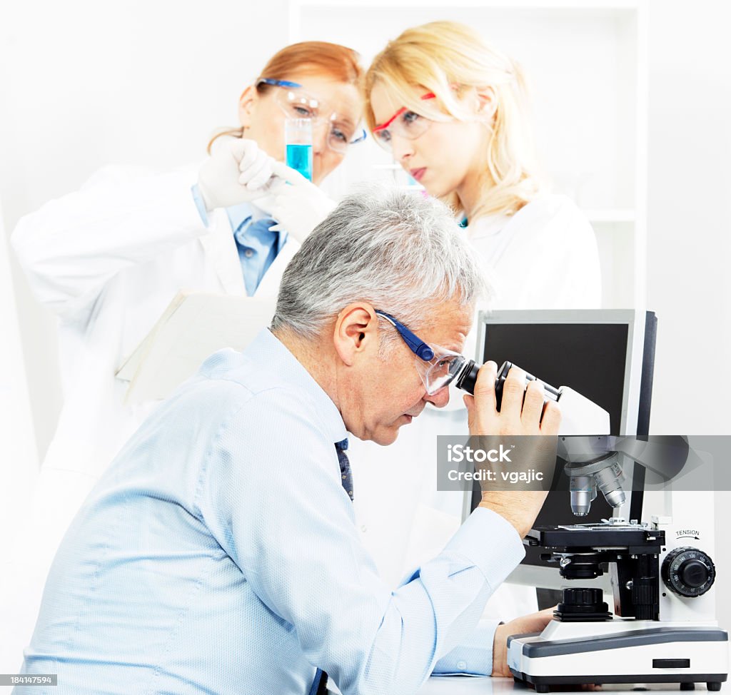 Group of scientists in a laboratory Senior male scientist researcher looking samples under stereo microscope and two female scientists preparing samples in background, selective focus to senior scientist Active Seniors Stock Photo