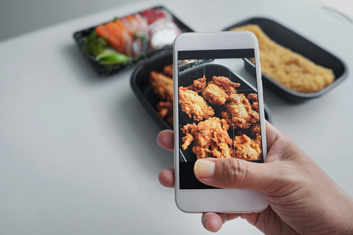 Close-up POV shot unrecognizable female influencer hand holding smartphone taking pictures of a takeout meal. There's a Japanese Fried Chicken (Karaage Chicken), fresh sashimi and chicken katsu.