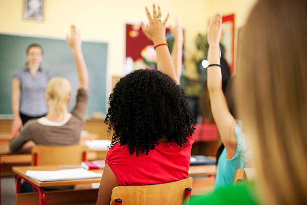 Students at the classroom answering questions. Rear view of students with raised hands. The focus is on the girl with black hair. teenage high school girl raising hand during class stock pictures, royalty-free photos & images