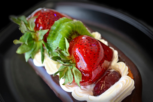 Glazed fruit strawberry and Kiwi on whip cream chocolate covered eclair on dark plate and black background.