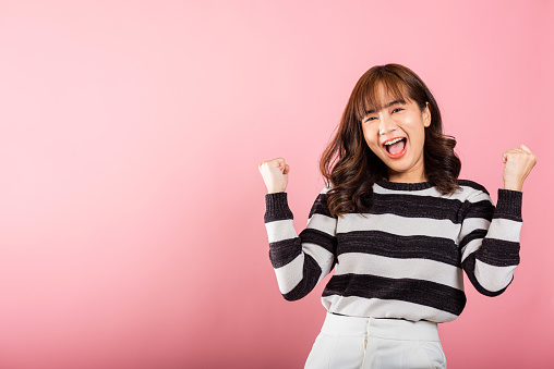 Portrait of a jubilant Asian woman with raised fists, celebrating her success with a happy expression. Studio shot isolated on a pink background, conveying victory and joy.
