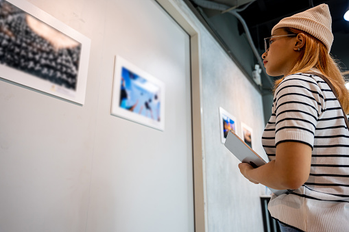 Asian woman holding tablet at art gallery collection in front framed paintings pictures on white wall, Young person at photo frame hold digital book leaning against at show exhibition artwork gallery