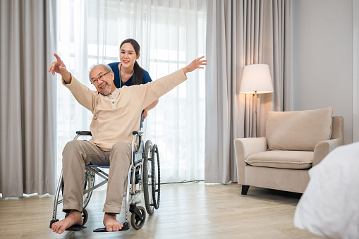 Happy curator person doctor pushing wheelchair and run elderly disabled patient freedom raising arm at hospital, senior retired man sitting on wheelchair having fun with young woman nurse, health care