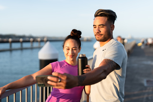 Happy Pacific Islander couple in casual clothing taking selfie using smart phone outdoors.