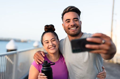 Happy Pacific Islander couple in casual clothing taking selfie using smart phone outdoors.