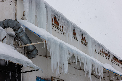 Multiple frozen winter icicle stalactites hanging over the edge of the overflowing roof gutters along the eaves and drainage downspouts of a suburban Rochester, New York State residential district house.