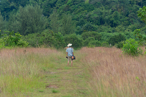 An Asian adult man wearing hat carrying camera and tripod walking in savannah field at giant cliff, Hat Thai Mueng beach, Phangnga province. Sustainability travel concept.