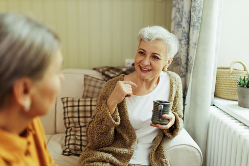 Stylish Caucasian retired female in warm cardigan holding mug looking at her unrecognizable female friend having curious facial expression, listening attentively. Two elderly women talking