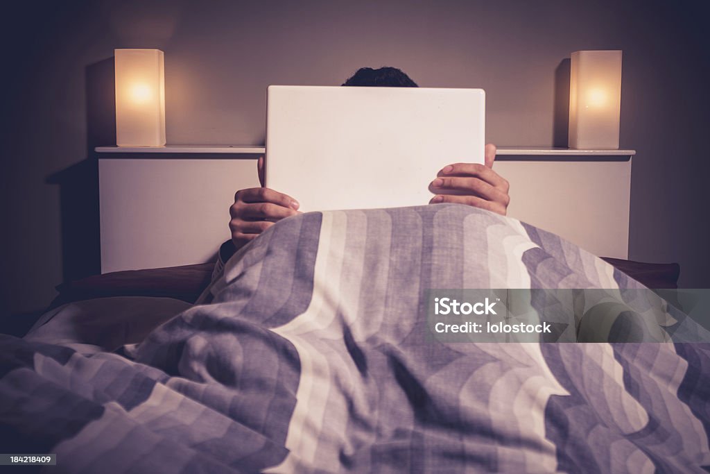 Man is sitting in bed behind his laptop Man is sitting in bed at night under cover and behind his laptop, only his hands and the top of his head is visible Adult Stock Photo