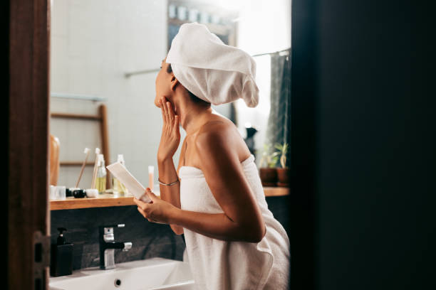 una hermosa mujer envuelta en una toalla mirándose en el espejo mientras se aplica un poco de crema en la cara después de bañarse - facial cleanser fotografías e imágenes de stock