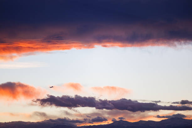 An airplane disappears into the distance as the golden light of the setting sun casts an orange glow on the dark clouds.