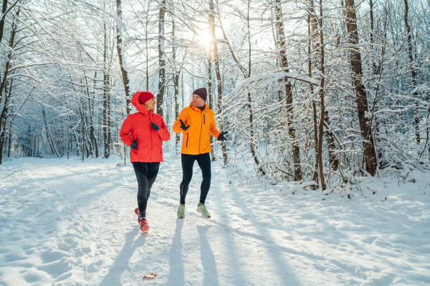 corridori sorridenti di mezza età della coppia di trail uomo e donna vestiti giacche antivento luminose che corrono parlando pittoresca foresta innevata durante la giornata gelida e soleggiata. persone sportive attive, immagine di allenamento invernale. - cold discussion outdoors snow foto e immagini stock