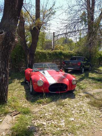 Remedios de Escalada, Argentina - Oct 8, 2023: Old red 1960s AC Shelby Cobra sport roadster replica under the trees at a classic car show in in a park.