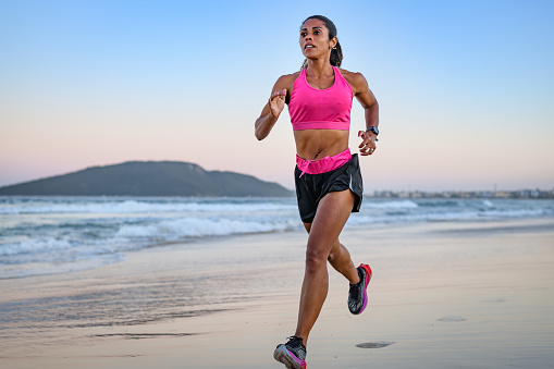 Black woman athlete doing training running drills on the beach at dusk in summer. Her focus and self-discipline will lead you to great sporting results at the finish line.