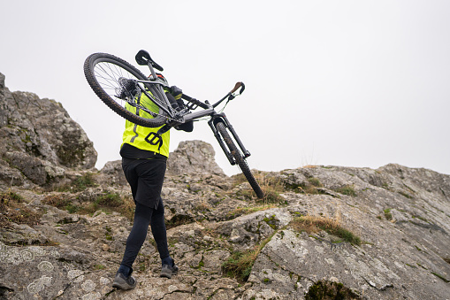 Man carrying mountain bike on his shoulder while moving up to the top of a rocky mountain on foggy day