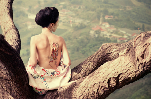 Asian style portrait of young woman with snake tattoo on her back sitting on the tree branch