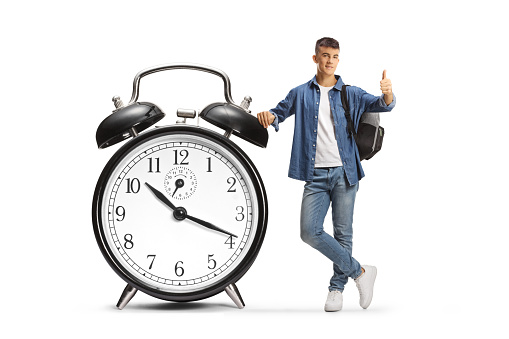 Male student leaning on a big alarm clock and gesturing thumbs up isolated on white background