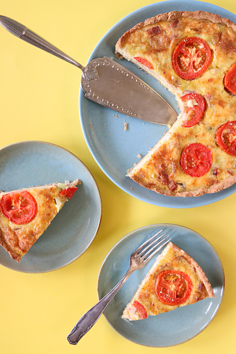 Stock photo showing close-up, elevated view of a fluted pastry crust, homemade, Quiche Lorraine, topped with tomato slices, on a blue plate against a yellow background.