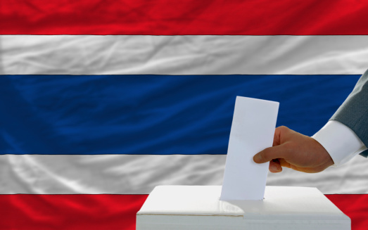 man putting ballot in a box during elections in thailand in front of flag