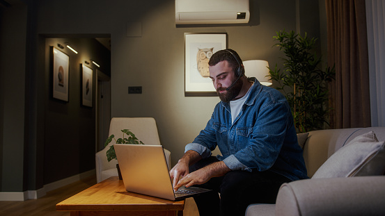 Bearded young adult business man wear wireless headset while sittin of sofa having video conference calling on laptop computer talk by webcam in online chat