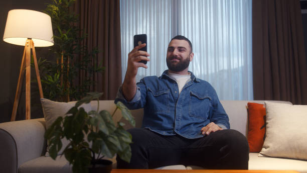 istock Young adult bearded man at home sitting on couch while having video call conversation through app with using smartphone, share news, talk to family living abroad, enjoy webchat virtual meeting 1842558446