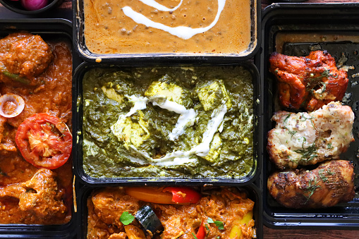 Stock photo showing close-up, elevated view of a selection of Indian takeaway dishes in black plastic food containers. Pictured is Chicken Bhuna, Palak paneer (cottage cheese and spinach puree), Dal makhani (black lentil curry), Murgh malai tikka (grilled chicken supreme) and Kadai (Kadhai) Chicken, named after the style of cooking pot used to make the recipe. Kadai Chicken is also known as Chicken Karahi.