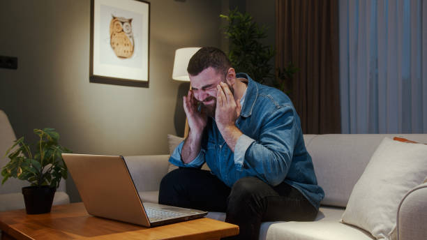 istock Young adult man sitting on sofa at home while using laptop having headache. Tired young man get having pain from a headache or migraine 1842572122
