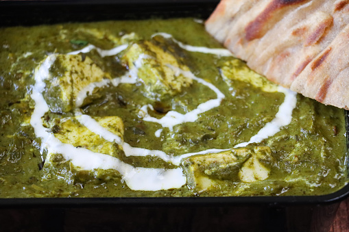 Stock photo showing close-up, elevated view of an Indian takeaway dish of Palak paneer (cottage cheese and spinach puree) garnished with a swirl of yoghurt served with lachha paratha (layered flatbread).