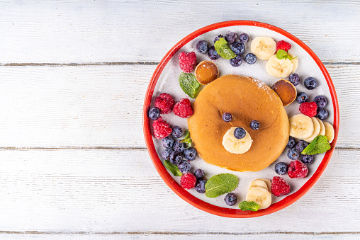 Funny and cute Christmas teddy bear pancakes on a red white plate decorated with fresh berry, fruit and powdered sugar, children's Christmas breakfast or brunch idea