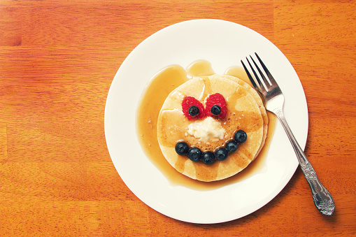 a plate of pancakes and syrup making a funny face with fruit.