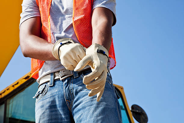 Workman at construction site putting on gloves Low angle, cropped view of an African American young man putting on work gloves.  He is a construciton worker, wearing an orange safety vest and jeans.  Part of the crane is visible behind him, against the clear, blue sky. crane machinery photos stock pictures, royalty-free photos & images