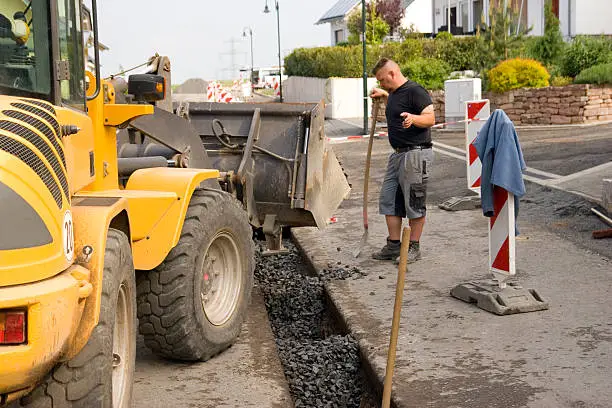 "+Focus on loader+ Shot of a wheel loader and a road worker at a construction site, road worker giving hand signals for tilting of the gravel in to the ditch. Drainage pipes were laid in a ditch, the trench is backfilled with gravel and then sealed with asphalt."