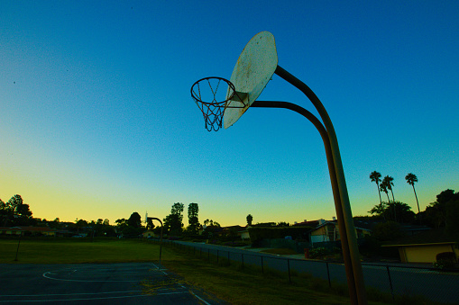 A playground basketball hoop and backboard stands against a dramatic sky at sunrise or sundown. The image evokes the timelessness of youth sports, the importance of sports in western culture, and the solitary pursuit of athletic excellence.