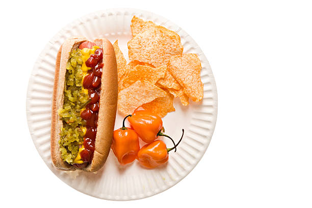 One Hot Hotdog "An overhead close up of a white paper plate containing a white bun with hotdog dressed with green relish,mustard and ketchup, a pile of spicy potato chips and three ripe habanero peppers for the pepper lovers  or the unwary... Isolated on white." relish green food isolated stock pictures, royalty-free photos & images