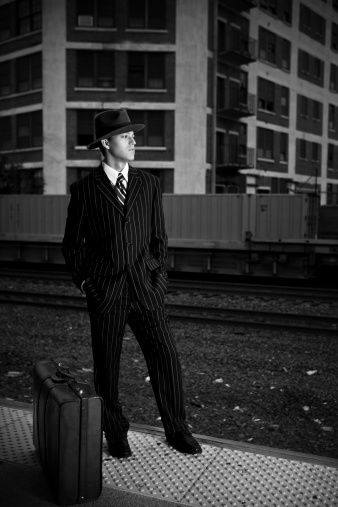 A man dressed in 1940s style waiting on a train platform with a suitcase