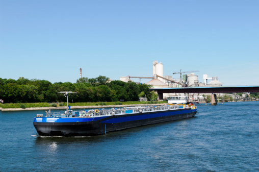 A river barge is traveling down the Rhine River carrying a cargo of fuel.  A factory can be seen in the background on the river shore.