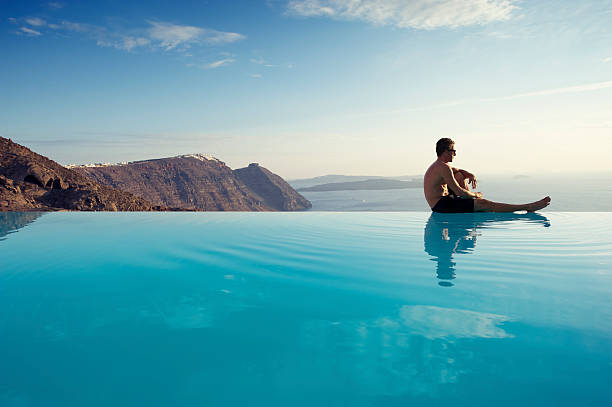 Young Man Sitting Infinity Pool Edge Caldera View Santorini Greece Man sits on the edge of an infinity pool gazing out at the misty Mediterranean seascape infinity pool stock pictures, royalty-free photos & images