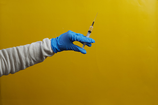Hand in medical glove holding syringe ready to inject on yellow background