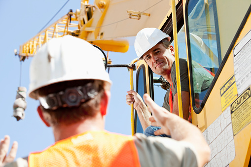 Low angle view of two construction workers operating a crane.  The driver is looking down at a man on the ground who is talking to him, giving directions and gesturing with his hands.  They are both wearing white hard hats and orange safety vests.  The focus is on the man in the cab of the construction equipment and we see the back of the head of the other man who is maybe the foreman.