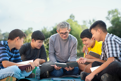 Group of asian family spending time vacation in local park by reading books, talking a joke story to each other happily, soft focus.