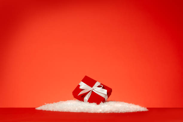 confezione regalo rossa festiva con fiocco bianco sdraiato nella neve su sfondo rosso. concetti di celebrazione del natale o delle festività. - fake snow foto e immagini stock