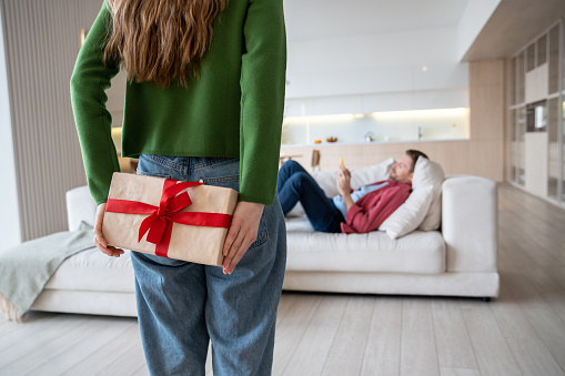 Woman hiding unexpected surprise giftbox braced with red ribbon, covered with festive paper, behind back. Female going to congratulate man lying on sofa with anniversary, birthday, St Valentine day