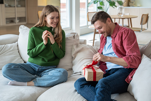 Thrilled intrigued man unbracing red ribbon, trying to unwrap cardboard giftbox. Pleasantly smiling merry loving woman looking with excitement and hope that beloved darling man will like present